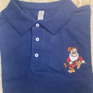 May include: A navy blue polo shirt with a white embroidered design of a dwarf holding a golf club. The shirt has a button-up collar and a button placket.