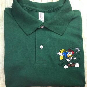 Op de afbeelding: Een groen poloshirt met een geborduurd beeld van Donald Duck die golf speelt. Het shirt heeft een knoopsluiting en een Jerzees-merklabel.