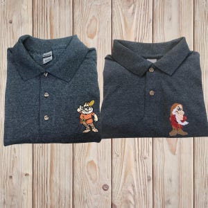 Op de afbeelding: Twee donkergrijze poloshirts met geborduurde afbeeldingen van de dwerg Doc uit Sneeuwwitje. De dwerg links draagt een bril en kijkt naar rechts. De dwerg rechts kijkt naar links.
