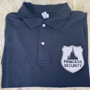 Może przedstawiać: Koszulka polo w ciemnoszarym kolorze z naszywką haftowaną na biało po prawej stronie. Naszywka przedstawia czarny zamek i napis "Princess Security".