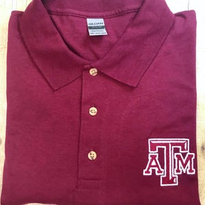 Puede incluir: Polo de color burdeos con una "T" y una "AM" bordadas en blanco en la manga derecha. La camisa está hecha por Gildan.