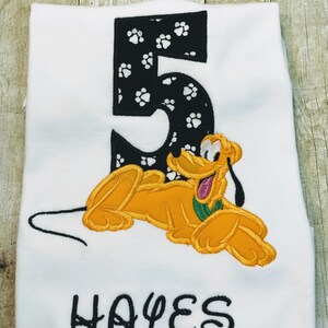 Puede incluir: Camiseta blanca con un número 5 negro con huellas de pata blancas, un perro de dibujos animados amarillo bordado y el nombre "HAYES" en negro.