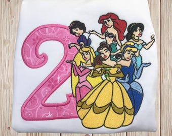 May include: Pink embroidered number two with seven Disney princesses, including Snow White, Ariel, Belle, Cinderella, Aurora, and two others. The princesses are all wearing their signature dresses.