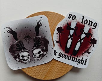 May include: Two sticker designs. One features two black and white skull figures wearing military hats with a single eye symbol above them. The other sticker features two black ballet shoes with red roses and the text "so long & goodnight".