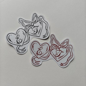 May include: Two heart-shaped stickers. One is black and white, the other is red and white. Each heart has the letter "E" inside. Ribbons and bows are also featured in the design.