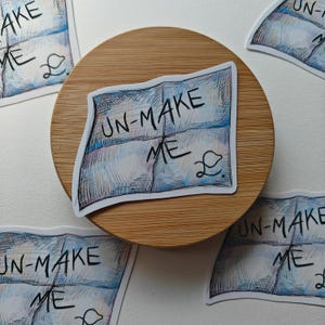 May include: A collection of stickers with the text "UN-MAKE ME" in a handwritten style. The stickers feature a blue and white watercolor design and a small, stylized drawing. They are arranged on a wooden coaster.