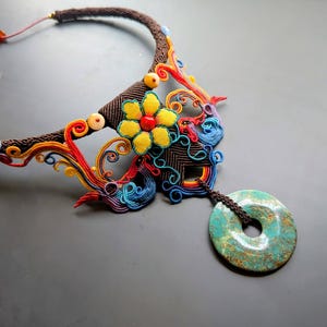 May include: A handcrafted necklace featuring a brown braided collar with colorful floral and swirl accents. A turquoise-colored circular pendant hangs from the necklace. The necklace has a unique, artistic design.