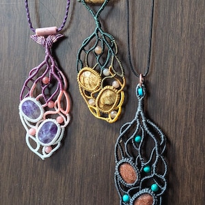 May include: Three handmade macrame necklaces with different colored stones and cords. The necklaces feature a leaf-shaped design with a knotted pattern. The stones are amethyst, sunstone, and carnelian.