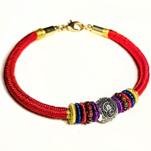 May include: Red cord bracelet with gold-colored accents and a silver-colored centerpiece. The bracelet features a series of colorful beads in blue, purple, and brown. The clasp is gold-colored.