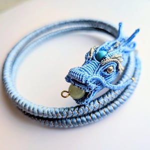 Puede incluir: Un brazalete de macramé azul con un diseño de dragón. La cabeza del dragón está intrincadamente elaborada con hilos azules y plateados, acentuada con una piedra turquesa y una cuenta verde en la boca. El brazalete es de color azul claro.