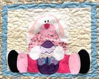 May include: A quilted Easter-themed wall hanging featuring a bunny holding a decorated egg. The bunny has pink and white accents, a floral patterned body, and black feet. The background is beige with a blue border.