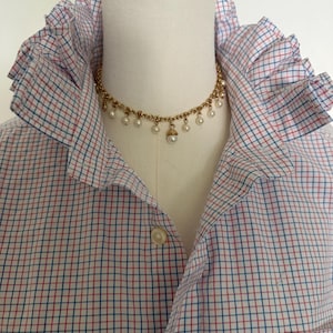 May include: A white button-down shirt featuring a red and blue grid pattern and a high ruffled collar. A gold-tone chain necklace with small dangling white pearls is worn around the neck of the mannequin displaying the shirt.