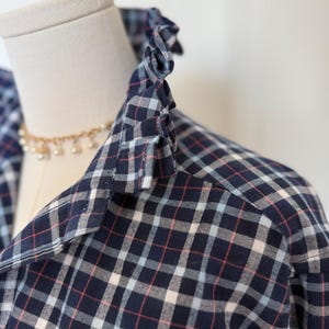 May include: A navy blue plaid shirt featuring a ruffled collar detail. The fabric pattern consists of intersecting lines in shades of light blue, white, and thin red, creating a classic checkered design across the garment's collar and shoulder area.