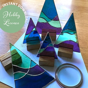 Pode incluir: Cinco enfeites de árvore de Natal de vidro colorido em tons de azul, verde e roxo. Cada enfeite é montado em uma base de madeira. Os enfeites estão dispostos em uma superfície branca com um rolo de fita de cobre.