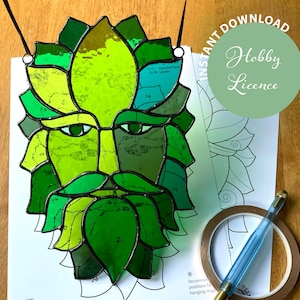 Op de afbeelding: Een glas-in-loodkunstwerk van een Green Man-gezicht in groentinten, met een baard en bladachtige details. Het kunstwerk wordt getoond op wit papier met een groene cirkel met de tekst "Instant Download Hobby Licence". Een blauwe tool en tape zijn ook zichtbaar.