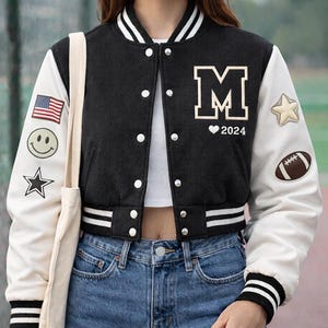 May include: Black and white varsity jacket with the letter "M" and "2024" on the front. The jacket has white sleeves with patches including a smiley face, star, American flag, star, and football. The jacket is worn with blue jeans.