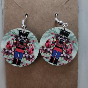 Può includere: Un paio di orecchini rotondi in legno con un design a forma di schiaccianoci. Gli orecchini sono bianchi con un motivo floreale verde e uno schiaccianoci rosso e blu al centro. Gli orecchini hanno ganci d'argento.