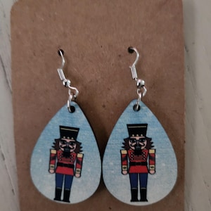 Può includere: Un paio di orecchini in legno a forma di goccia con sfondo blu e un'immagine stampata di uno schiaccianoci in uniforme rosso e blu. Gli orecchini hanno ganci d'argento.