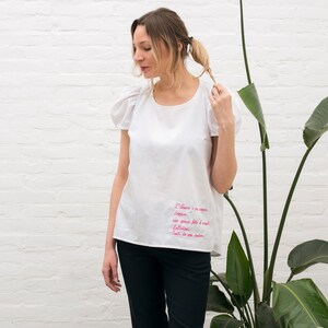 Peut inclure: Un chemisier blanc à manches courtes avec une citation brodée en rose en français. La citation dit "J'aime a me promener, voyager, avec vous faire l'oeil, Etre libre, Tout ce que je veux".