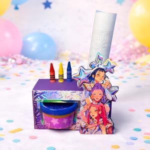 May include: A purple box with a blue and purple container, three crayons, and a decorative paper roll. A holographic sticker of three anime-style characters is attached to the box. Balloons and confetti are in the background.