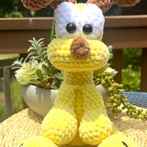 May include: A handmade, plush yellow dog toy with brown ears, a white face, and black eyes. The dog has yellow legs and round feet. The toy is sitting on a woven mat, with greenery in the background.