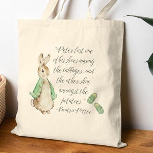 May include: Cream-colored tote bag featuring a watercolor illustration of Peter Rabbit in a green jacket. The bag includes a quote from Beatrix Potter: "Peter lost one of his shoes among the cabbages, and the other shoe amongst the potatoes."