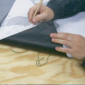 May include: A person's hand is tracing a design on a piece of wood with a pencil. A black sheet of paper is being peeled back to reveal the design.