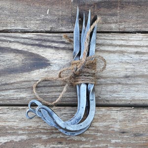 May include: A set of three hand-forged metal forks with a dark grey finish, featuring twisted handles and curled ends. The forks are bundled together and secured with a piece of natural brown jute twine tied in a knot around the center of the handles.