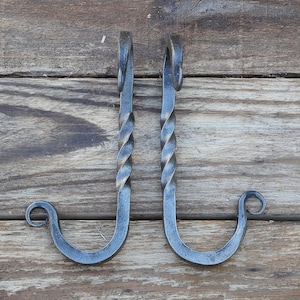 May include: Two dark gray metal hooks featuring a twisted vertical shaft and a curved bottom end with a small decorative scroll. Each hook has a loop at the top for mounting, showcasing a textured, hand-forged appearance with metallic highlights.