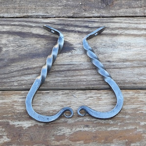 May include: Two hand-forged iron hooks with a dark grey metallic finish. Each hook features a twisted vertical shank that transitions into a curved J-shape at the bottom and includes a flat mounting tab with a pre-drilled hole at the top end.