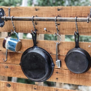 May include: A wall-mounted pot rack made of a horizontal wooden bar with four hand-forged metal S-hooks. A blue and brown ceramic mug, two black cast iron skillets, and a metal kitchen tool hang from the hooks. A black sign reads Pot Rack Hand Forged.