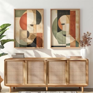 Könnte beinhalten: Zwei gerahmte abstrakte Kunstdrucke mit geometrischen Formen in Orange-, Beige-, Schwarz- und Grüntönen. Darunter ein helles Holzschrank mit gewebten Paneelen und eine dekorative Pflanze in einem gewebten Korb.