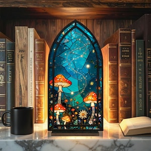 May include: A stained glass panel featuring a whimsical mushroom design with orange caps and white spots, set against a blue night sky with twinkling lights. The panel is framed in black and sits on a wooden base, with a black mug and books in the background.