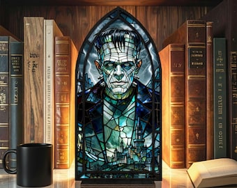 Puede incluir: Un panel de vidrieras con el monstruo de Frankenstein en un diseño de arco gótico. La obra de arte utiliza tonos de azul, verde y negro, con una escena de castillo en la parte inferior. El panel se exhibe en un estante con libros y una taza negra.