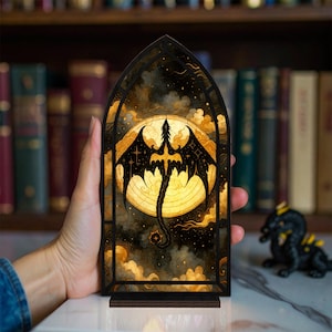 May include: A decorative wooden panel featuring a dragon design. The dragon is black with outstretched wings, set against a yellow moon and a cloudy, dark background. The panel is in a stained-glass style, with a wooden base.