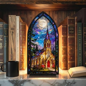 May include: A stained glass church scene with a full moon and starry sky. The church has a red door and steeple. The artwork is displayed on a shelf with books and a black mug.