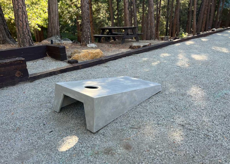 May include: A gray concrete cornhole board with a round hole in the center. The board is sitting on a gravel surface.