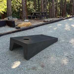 May include: A black, angled, wooden bean bag game board with a hole in the center. The board is sitting on a gravel surface.