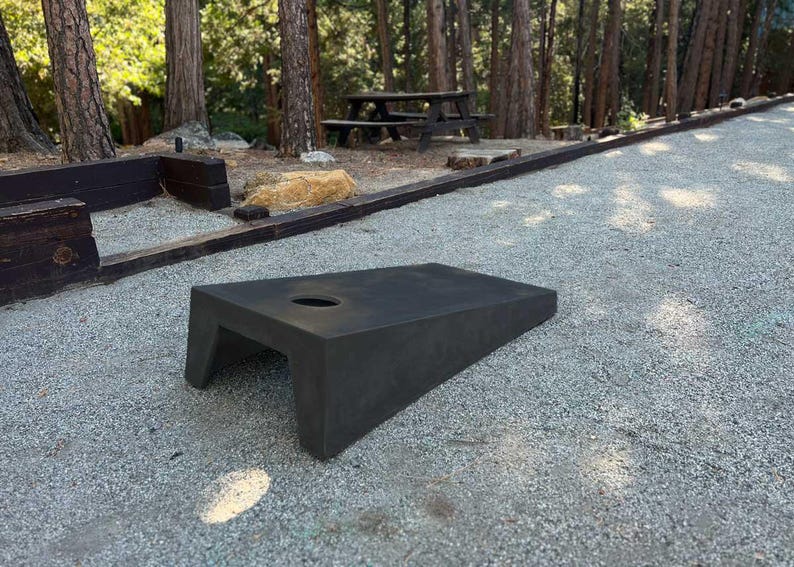 May include: A black, angled, wooden bean bag game board with a hole in the center. The board is sitting on a gravel surface.