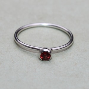 May include: A simple silver band ring with a small, round, red gemstone.