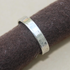 May include: A silver ring with a hammered texture. The ring is a simple band design, reflecting light. The ring is displayed on a brown, textured surface. The ring is a piece of jewelry.