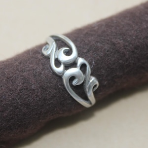 May include: A silver ring with an openwork design featuring swirling, scroll-like shapes. The ring is displayed on a dark brown, textured surface, highlighting its intricate details and metallic sheen. The ring's design is symmetrical and elegant.
