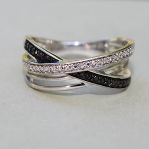Può includere: Un anello argentato con un design a incrocio. Una fascia è incastonata con piccole pietre chiare, mentre un'altra presenta pietre nere. L'anello ha una finitura lucida ed è posto su uno sfondo chiaro.