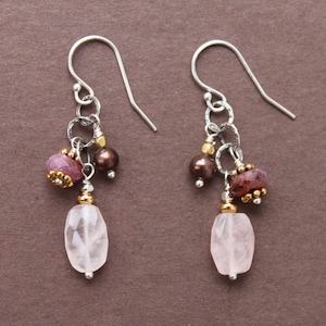 Könnte beinhalten: Ohrringe zum Hängen mit silberfarbenen Haken. Jeder Ohrring hat einen facettierten, hellrosa, ovalen Stein, ergänzt durch kleine, runde, dunkelbraune Perlen und eine Ansammlung von rötlich-rosa und goldfarbenen Perlen.