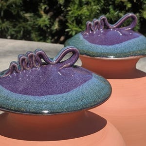 Puede incluir: Dos jarrones de cer&aacute;mica color terracota con tapas a juego. Las tapas son de color p&uacute;rpura intenso con un borde verde azulado y un asa decorativa. Los jarrones se exhiben al aire libre con luz natural.