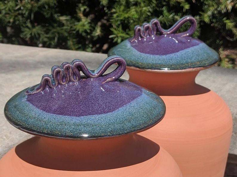 Puede incluir: Dos jarrones de cer&aacute;mica color terracota con tapas a juego. Las tapas son de color p&uacute;rpura intenso con un borde verde azulado y un asa decorativa. Los jarrones se exhiben al aire libre con luz natural.
