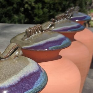 Puede incluir: Una fila de jarras de cer&aacute;mica de color terracota con tapas a juego. Las tapas est&aacute;n esmaltadas con una mezcla de colores, incluyendo verde, morado y azul. Cada tapa tiene un dise&ntilde;o de asa &uacute;nico. Las jarras est&aacute;n dispuestas en l&iacute;nea.