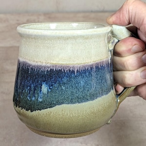 May include: A ceramic mug featuring a wide, rounded body and a sturdy handle. The upper portion is a creamy off-white, transitioning into a deep blue and purple drip glaze in the center, while the base is a textured, earthy tan color.