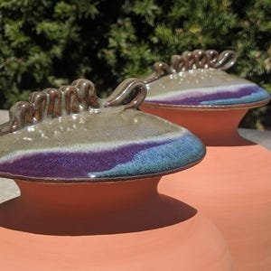 Puede incluir: Dos jarrones de cer&aacute;mica color terracota con tapas a juego. Las tapas presentan tonos verdes, morados y azules, con un asa marr&oacute;n texturizada. Los jarrones son probablemente para fines decorativos.