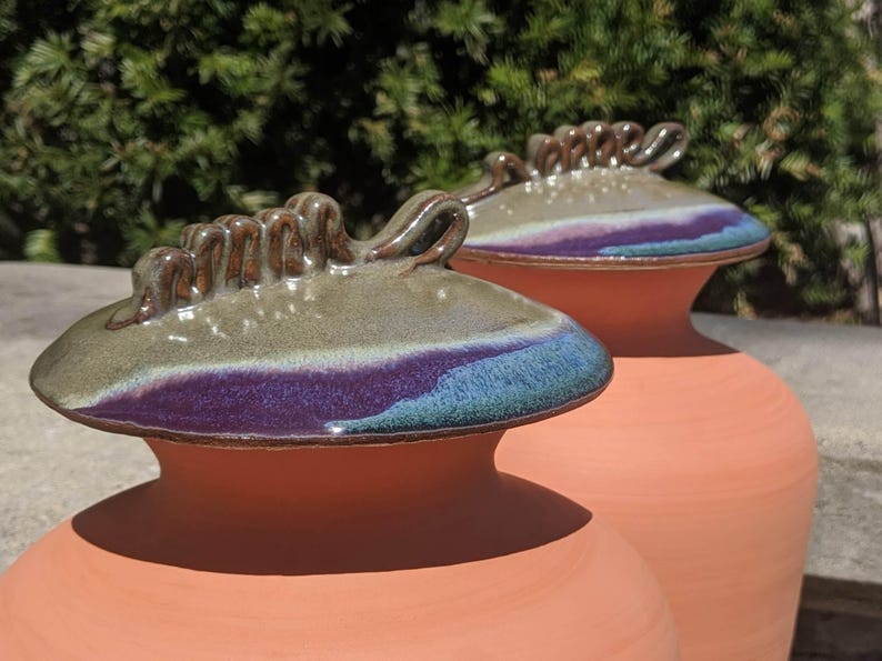 Puede incluir: Dos jarrones de cer&aacute;mica color terracota con tapas a juego. Las tapas presentan tonos verdes, morados y azules, con un asa marr&oacute;n texturizada. Los jarrones son probablemente para fines decorativos.